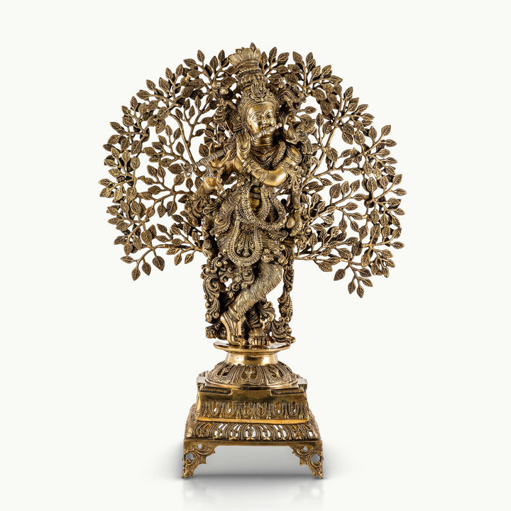 Antiqued brass Krishna statue playing the flute beneath a tree canopy.
