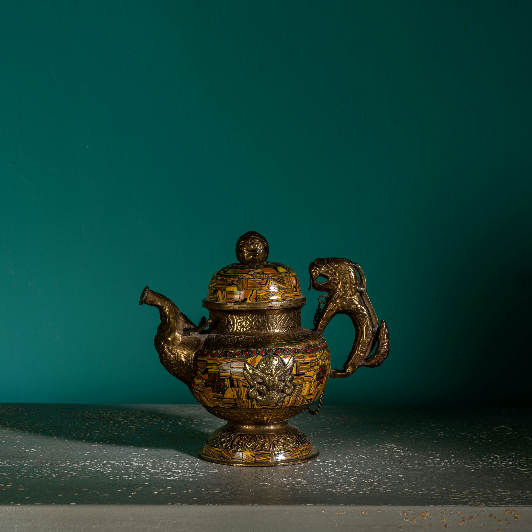 Vintage mosaic brass kettle displaying mythological guardians and a chained lid against a dark, antiqued metal background for a glass curio cabinet.