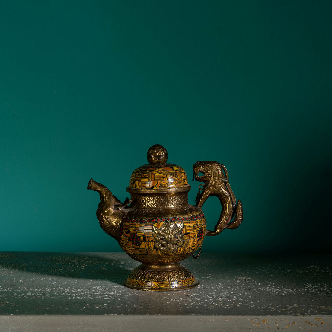 Vintage mosaic brass kettle displaying mythological guardians and a chained lid against a dark, antiqued metal background for a glass curio cabinet.