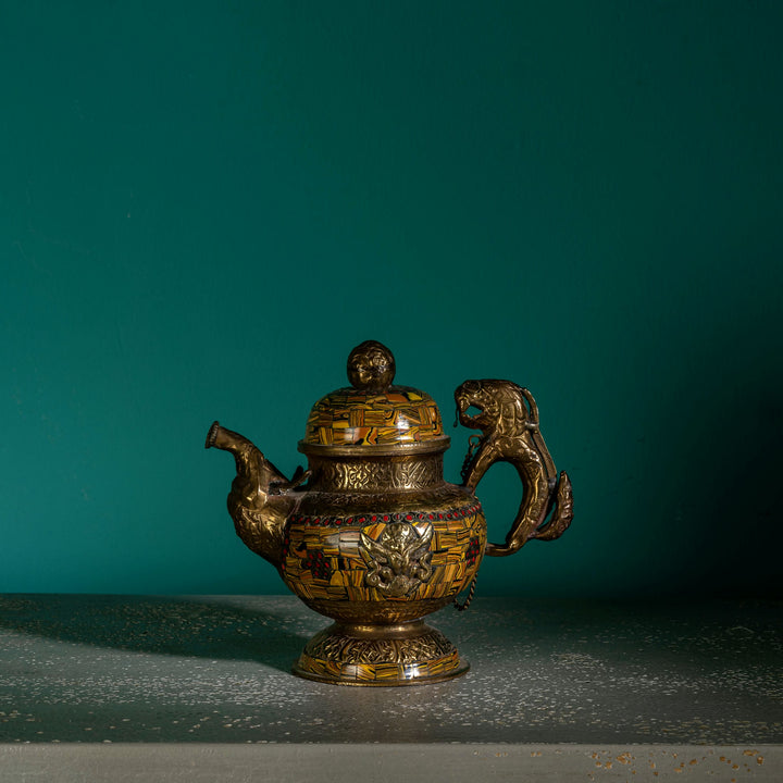 Vintage mosaic brass kettle displaying mythological guardians and a chained lid against a dark, antiqued metal background for a glass curio cabinet.