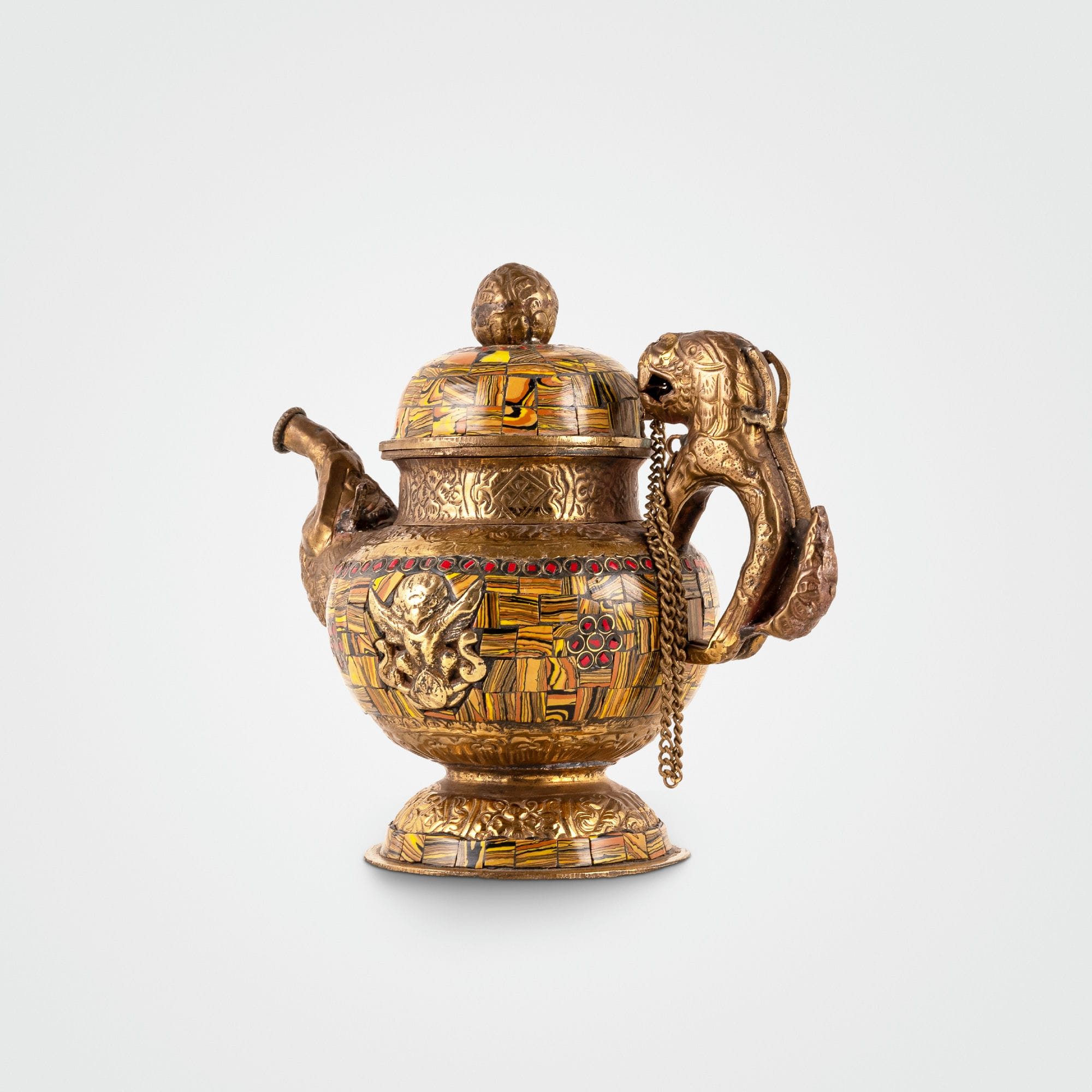 Vintage mosaic brass kettle displaying mythological guardians and a chained lid against a dark, antiqued metal background for a glass curio cabinet.