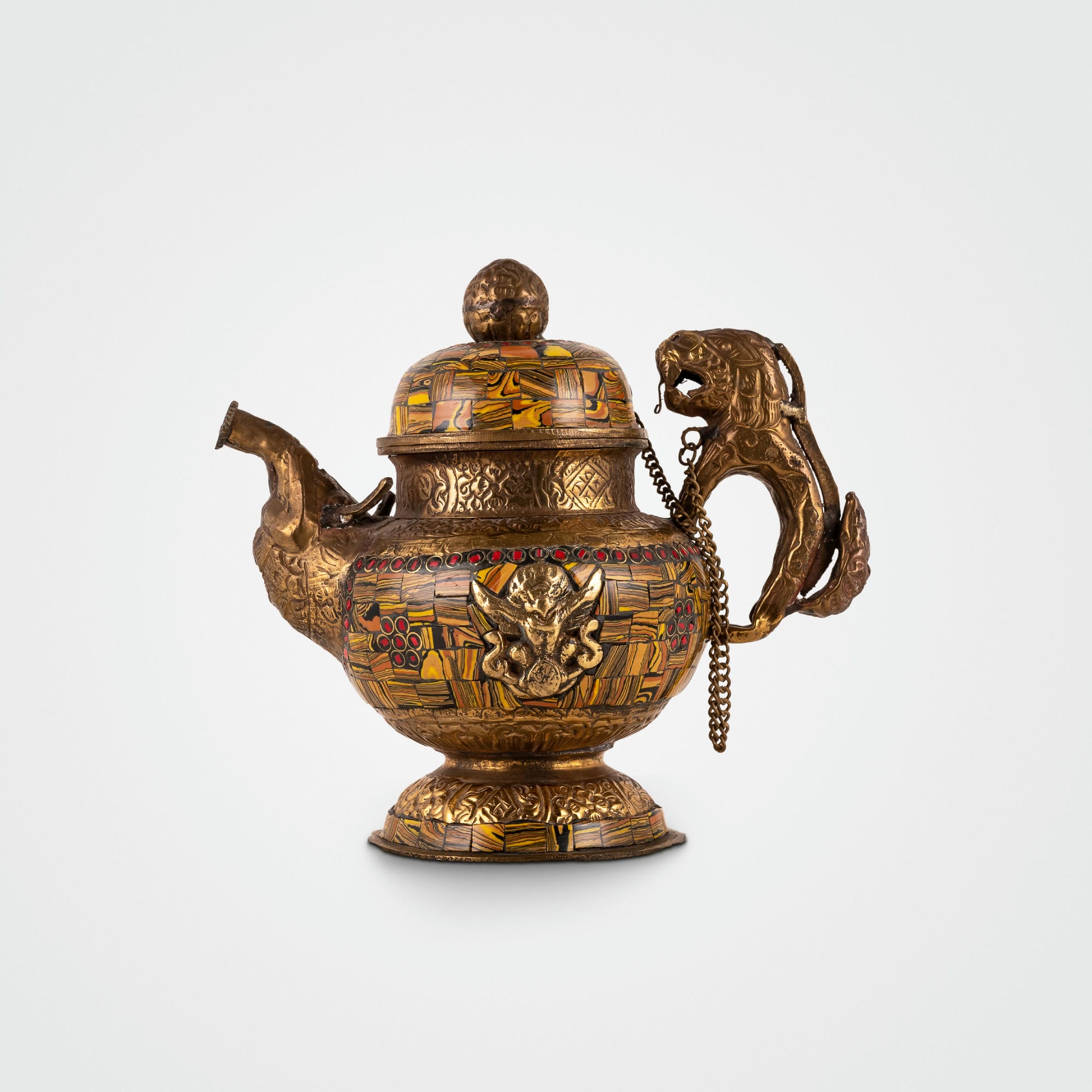 Stone chip inlay brass teapot featuring amber mosaics and a lion handle, positioned as a centerpiece for a glass curio cabinet. 