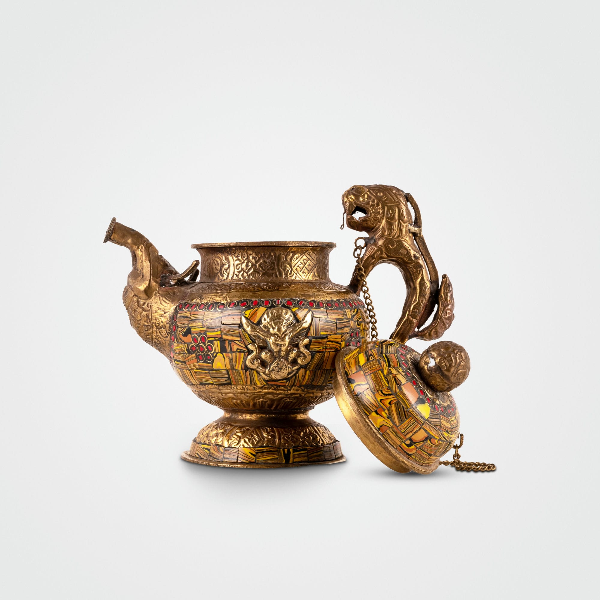 Vintage mosaic brass kettle displaying mythological guardians and a chained lid against a dark, antiqued metal background for a glass curio cabinet.